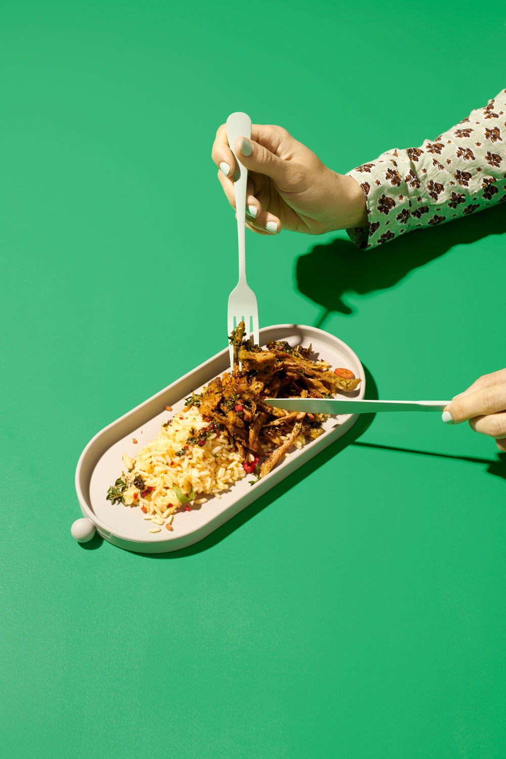 A hand using a fork and chopsticks to eat planted.pulled rice from an oval dish on a green background